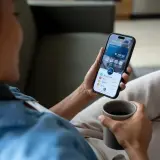 Kobieta z kubkiem kawy sprawdza na telefonie saldo konta bankowego. Aplikacja pokazuje dostępne środki i szczegóły wydatków.