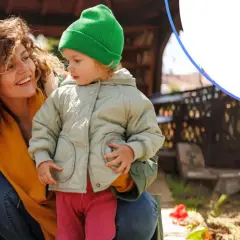 Uśmiechnięta mama z dzieckiem w ogrodzie. VeloBank lokaty na nowe środki to pewność przyszłości.