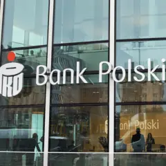 Logo PKO Bank Polski na szklanej fasadzie. Widać odbicia ludzi i miasta.