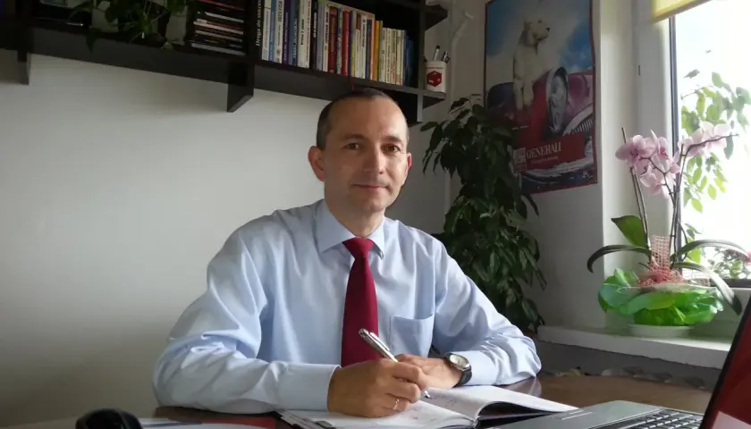 Mężczyzna w jasnej koszuli i czerwonym krawacie siedzi przy biurku, pisząc w notatniku.