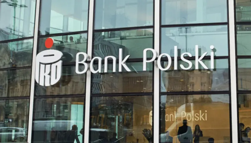 Logo PKO Bank Polski na szklanej fasadzie. Widać odbicia ludzi i miasta.